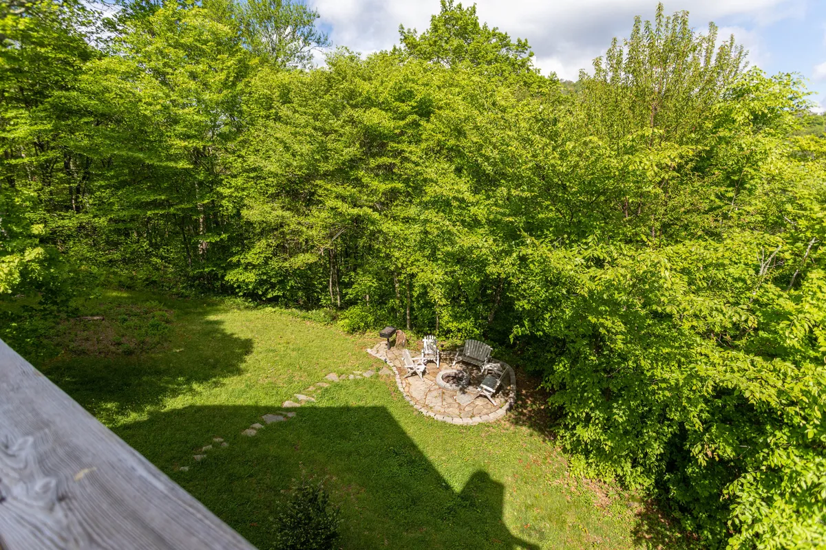 An inviting exterior seating area sits along a stone pathway, surrounded by mature trees. The atmosphere is serene and intimate, suggesting a perfect spot for quiet observation, reading, or gathering with friends while immersed in nature. The proximity to the woods and open space highlights easy access to outdoor activities and nature trails.
