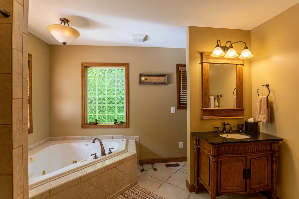 A serene bathroom with a jacuzzi tub set beside a window that offers a lush greenery view. A vanity with a mirror provides space for self-care, and the clean, bright ambiance invites relaxation and solitude.