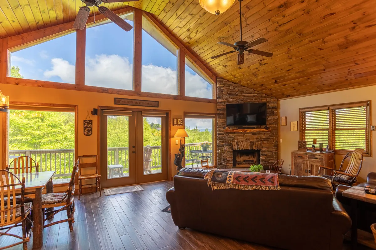 An open-concept dining and living space with a large wooden table set for family meals, comfortable seating, a prominent stone fireplace, and oversized windows offering lush views of the surrounding landscape.