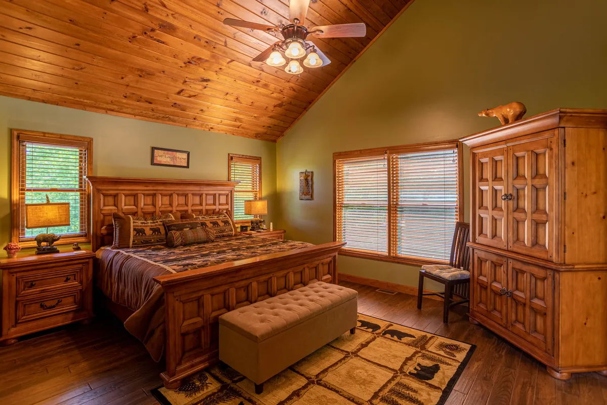 This image shows a cozy bedroom featuring a wooden bed frame adorned with decorative pillows, nightstands on each side, windows with blinds letting in natural light, and a rustic rug underfoot. The warm wood tones and soft textiles evoke a sense of relaxation and retreat.