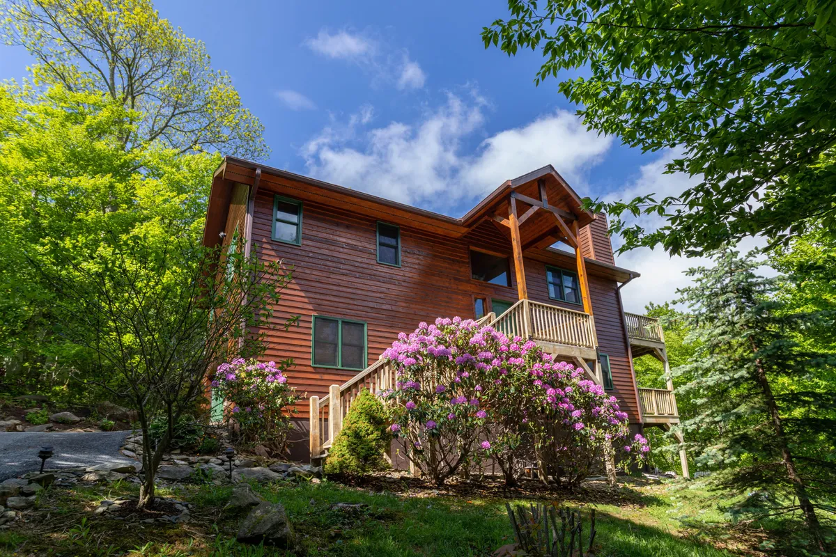 The image shows the wooden exterior of Bearadise Cabin surrounded by lush greenery and colorful, vibrant flowers in the foreground. The scene hints at ample outdoor space and includes the driveway and parking area leading up to the house, making it clear that parking for multiple vehicles is available.