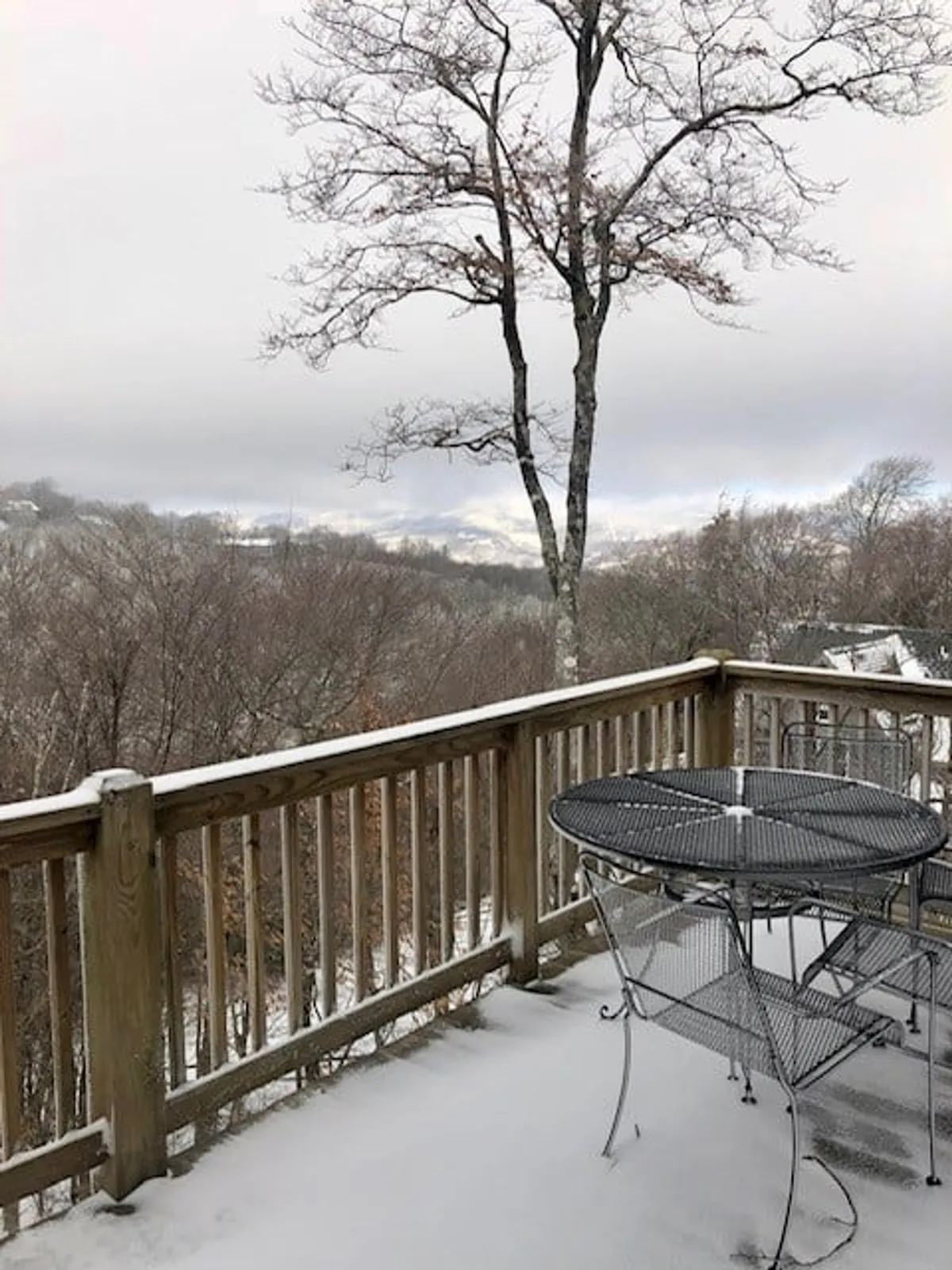 This image captures a serene winter scene: a snow-covered deck with a bare tree in the foreground and a sweeping mountainous landscape in the background. The deck, lightly dusted with snow, hints at a cozy space for enjoying the outdoors even in colder months.