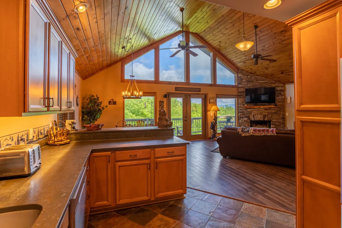 The image displays a bright interior with large windows overlooking surrounding greenery, a rustic wooden ceiling, a prominent stone fireplace, and a kitchen island. The scene depicts a seamless flow between living, dining, and kitchen areas, perfect for group gatherings and entertaining.