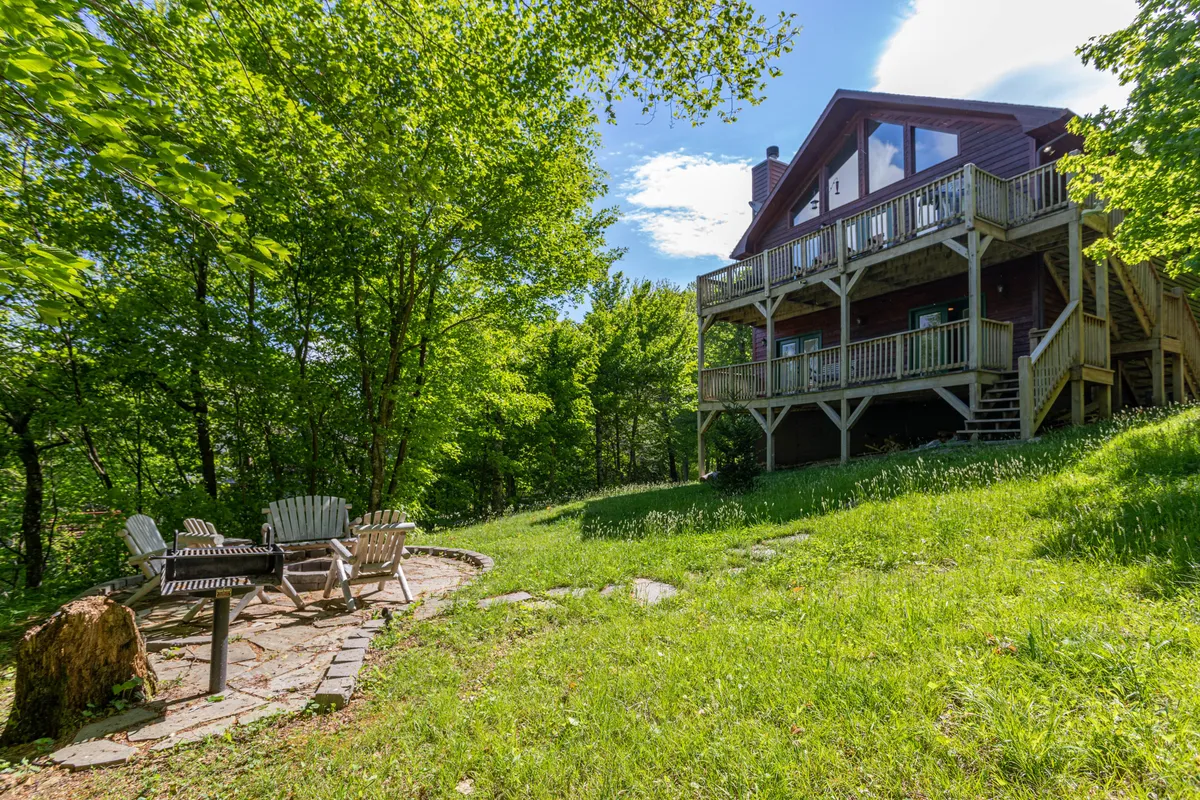 The image showcases the exterior of Bearadise Cabin on Sugar Mountain, with a charming balcony overlooking a lush green lawn, a spacious stone patio, and a cozy fire pit surrounded by seating. The setting is tranquil and inviting, ideal for outdoor gatherings, soaking in mountain views, or relaxing by the fire under the stars.