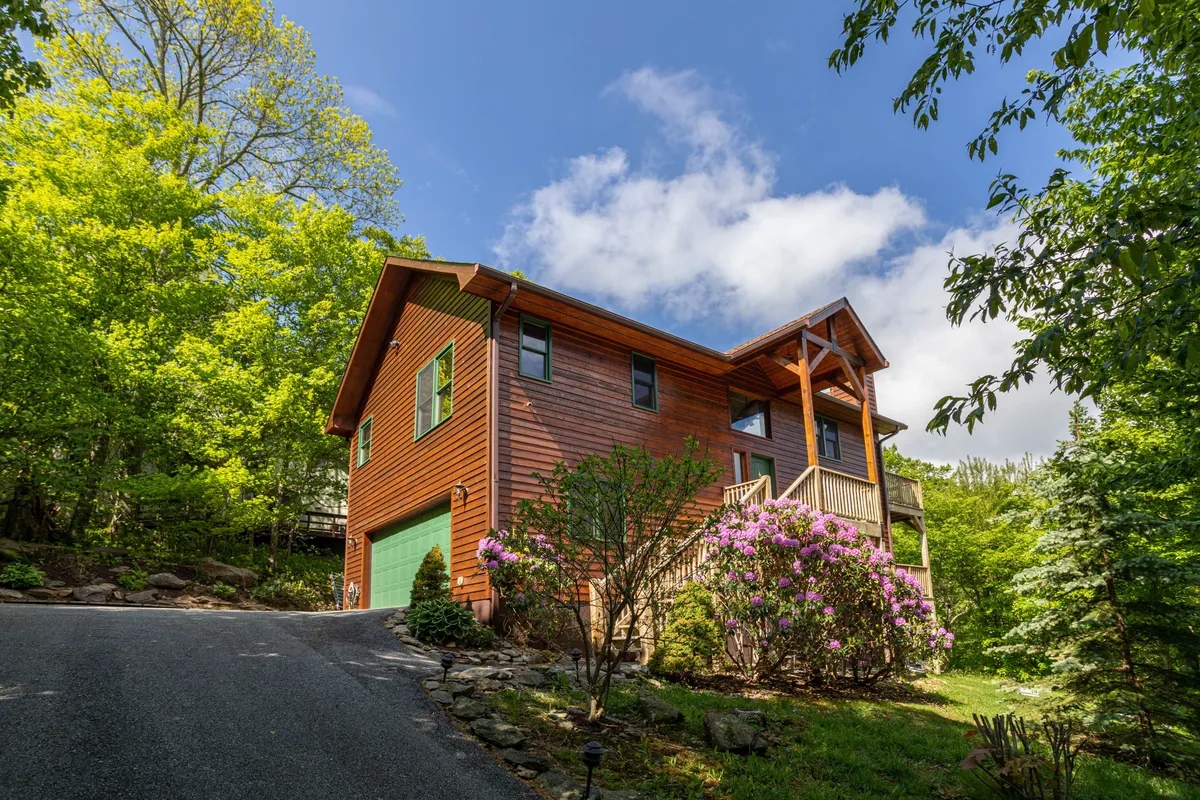 The image features the wooden house nestled among flowering bushes and lush green trees, with a clear blue sky overhead. The expansive yard and proximity to trails are implied, and the overall atmosphere is vibrant and welcoming.