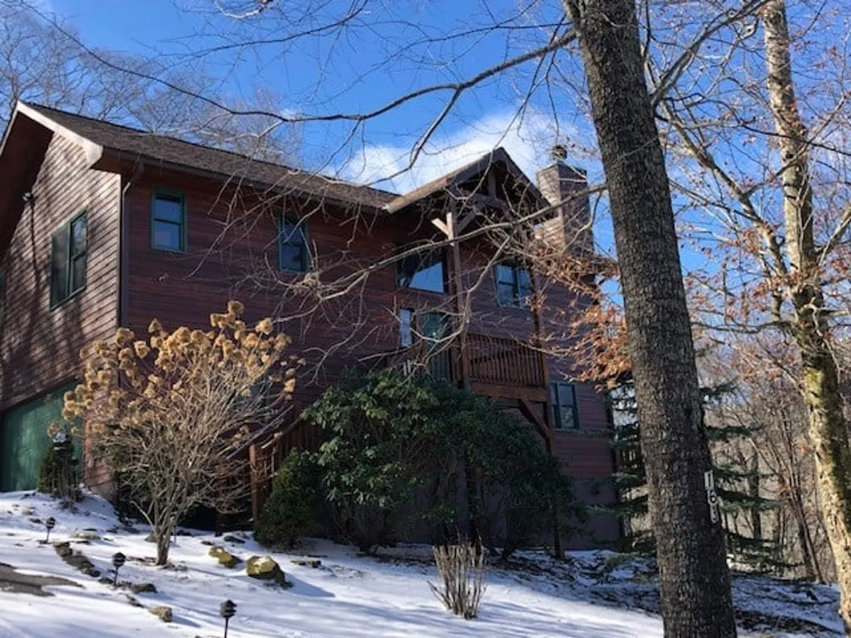 A cozy wooden cabin sits amidst a serene winter landscape, its front porch and the ground blanketed in fresh snow. Tall trees surround the property, and the porch offers a welcoming spot to watch the snowfall or prepare for a day of skiing. The scene captures the quintessential winter mountain getaway.