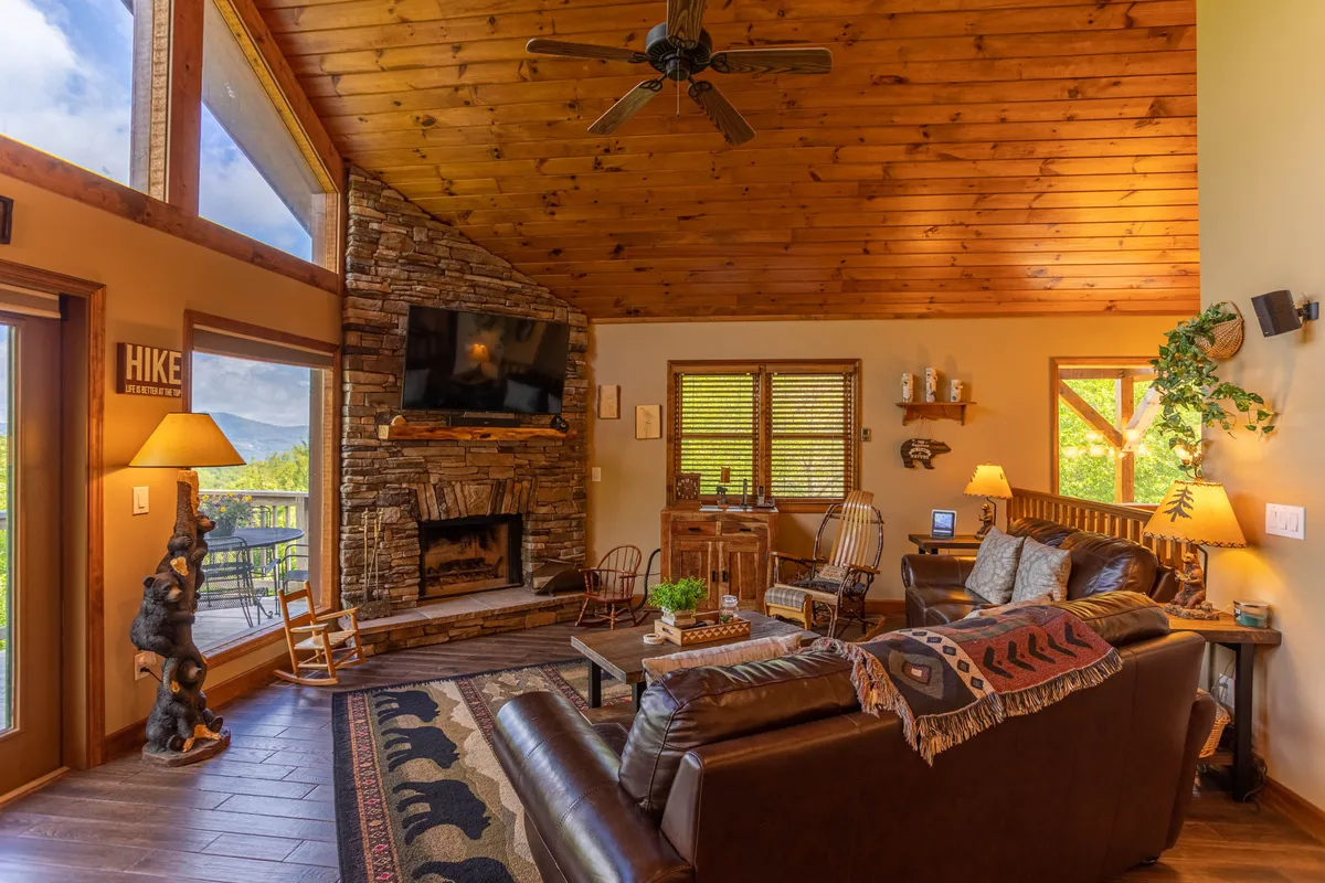 A cozy interior featuring a large stone fireplace, expansive windows that bring in natural light, a comfortable leather sofa, and a rustic wooden ceiling. Decorative plants add a homey touch, and the space is arranged to support group relaxation and easy living.