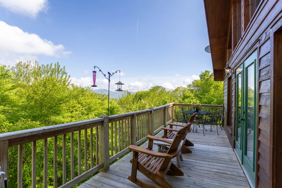 This image captures a wooden deck furnished with outdoor seating, a hummingbird feeder, and a breathtaking mountain view beyond the railing. The greenery and tranquil atmosphere invite relaxation and immersion in the natural setting.
