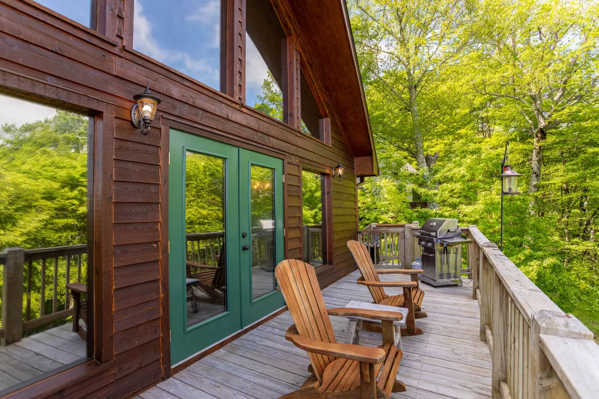 A spacious wooden deck extends from the home, surrounded by tall trees and lush greenery. Comfortable outdoor seating invites relaxation while the elevated position offers a partial view through the woods. The scene captures a sense of tranquility and direct access to nature, ideal for enjoying sunsets or watching wildlife from the comfort of the deck.